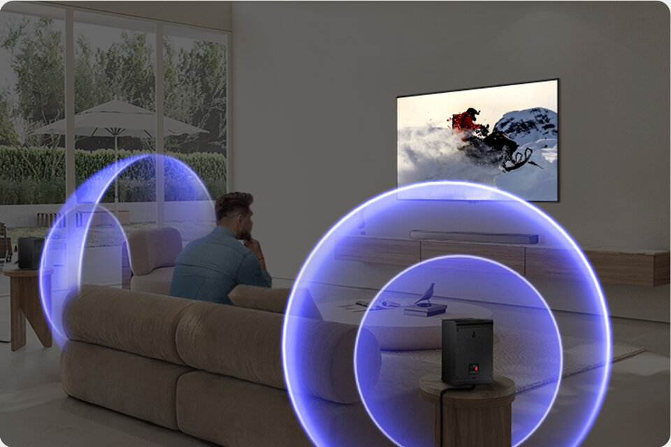 960c640 hw-b750fsound quality wireless surround sound compatible 2 W salonie znajduje się telewizor wyświetlający scenę z jazdą na nartach, a przed nim siedzi osoba na kanapie. Wokół soundbara i subwoofera widoczne są niebieskie fale dźwiękowe.