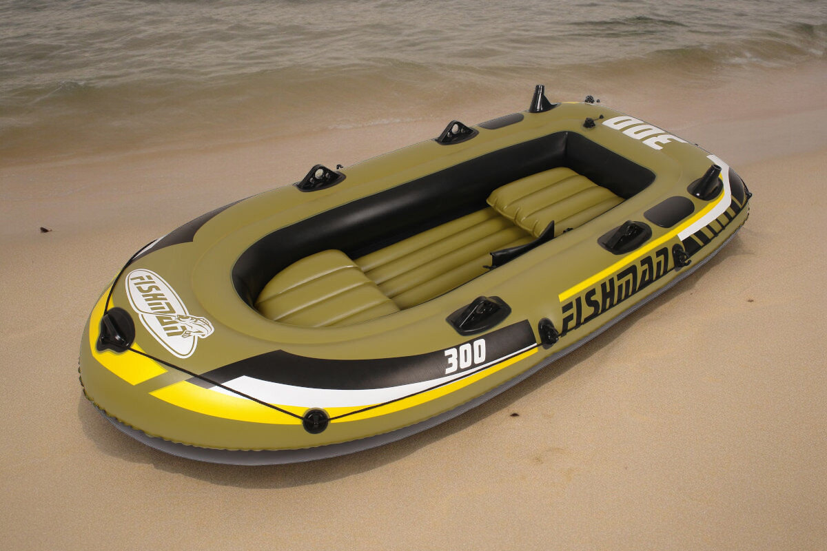 Ponton w kolorze oliwkowym z czarnymi i żółtymi akcentami leży na piaszczystej plaży. Na pontonie widoczne są napisy \'FISHMAN\' i \'300\'.