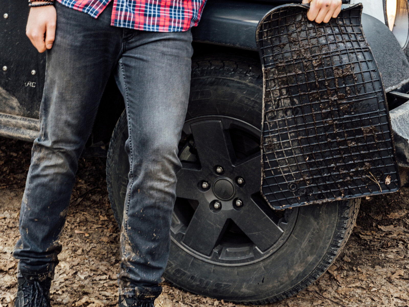 Osoba stoi obok samochodu terenowego, trzymając zabrudzoną gumową matę samochodową. W tle widoczne jest koło pojazdu z czarną felgą.