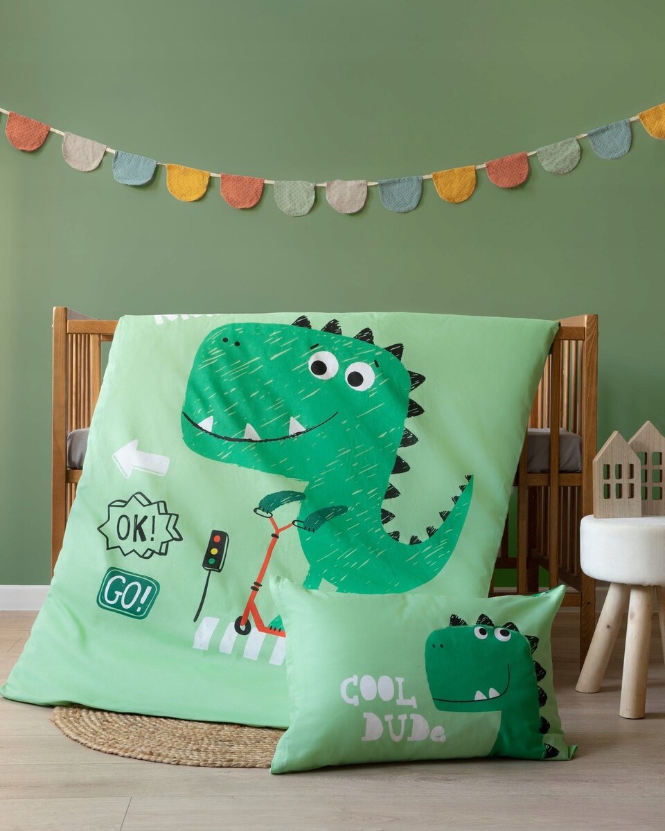 Na obrazie znajduje się pościel dziecięca z zielonym dinozaurem na hulajnodze oraz napisy \'OK!\', \'GO!\' i \'COOL DUDE\'. W tle widać drewniane łóżeczko i kolorową girlandę na zielonej ścianie.