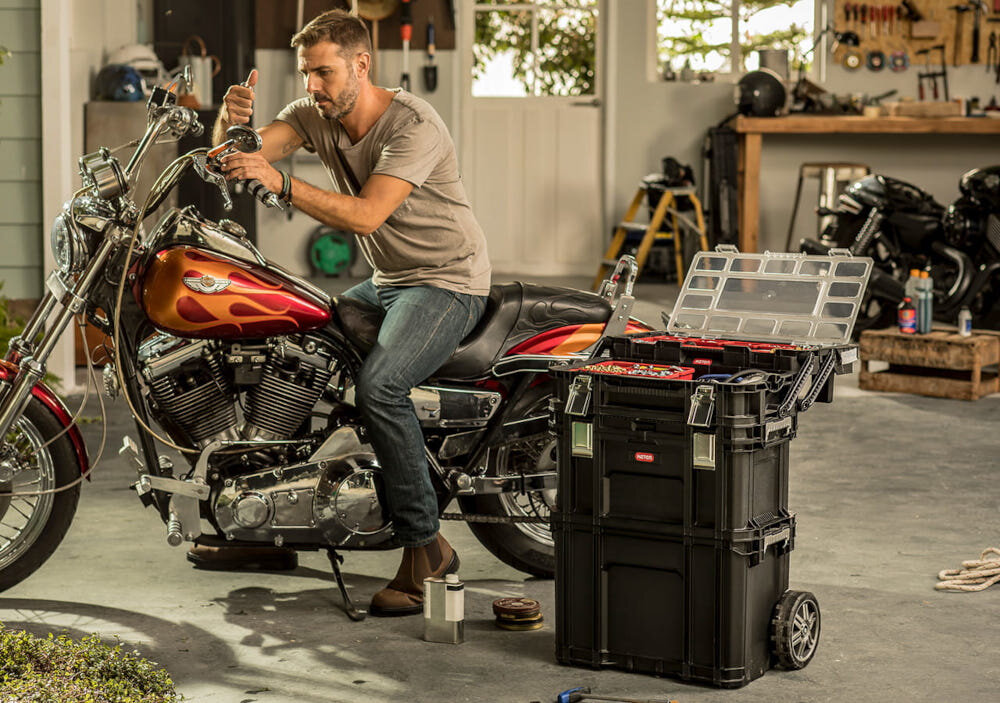 Mężczyzna pracuje przy motocyklu w garażu, obok znajduje się zestaw skrzynek narzędziowych na kółkach z otwartą pokrywą. Na skrzynkach widoczne jest logo \'KETER\'.