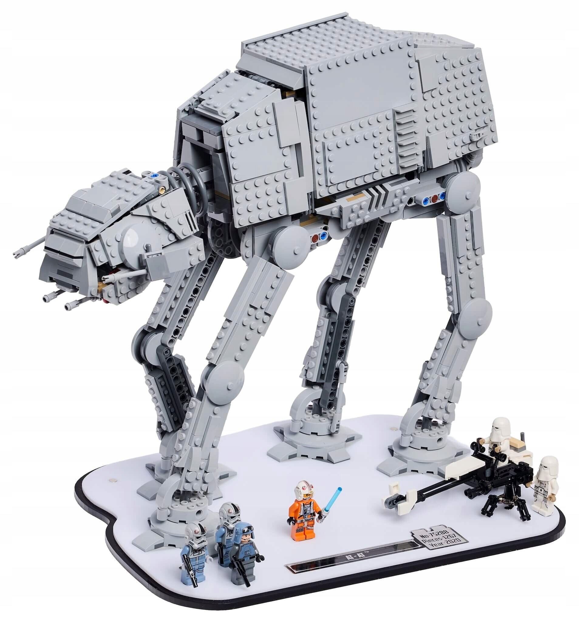 Model LEGO przedstawiający maszynę AT-AT z serii Star Wars, umieszczony na białej podstawie z kilkoma minifigurkami wokół. Na podstawie widoczna jest tabliczka z tekstem i symbolami.