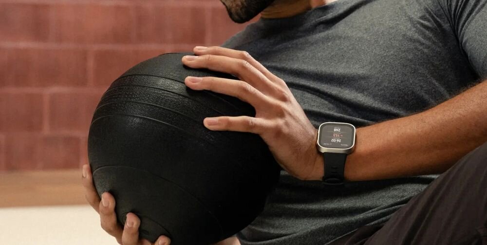 Osoba trzyma czarną piłkę do ćwiczeń, na nadgarstku ma smartwatch z wyświetlaczem pokazującym czas i datę. Smartwatch ma ciemny pasek i prostokątną tarczę.