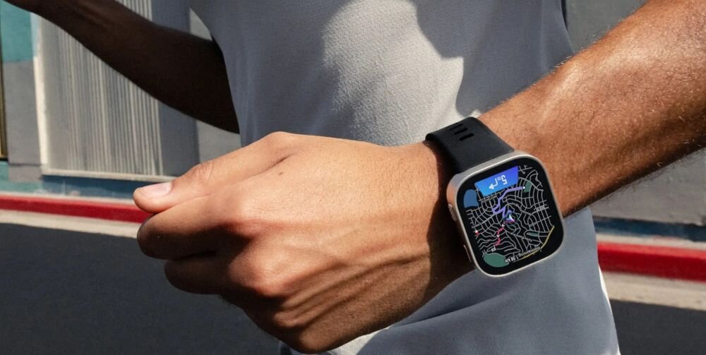 Osoba nosi smartwatch z ciemnoniebieskim paskiem, na ekranie widoczna jest mapa z trasą i ikoną GPS. W tle widać fragment ulicy i budynku.