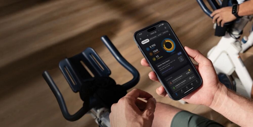 Osoba trzyma smartfon z aplikacją fitness, na ekranie widoczne są dane dotyczące aktywności fizycznej. W tle znajduje się rower treningowy.