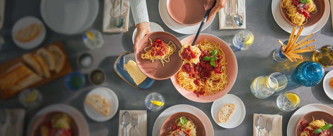 Stół zastawiony talerzami z makaronem spaghetti, na których znajduje się sos pomidorowy i liście bazylii. Wokół talerzy znajdują się szklanki z wodą i cytryną, sztućce oraz koszyk z pieczywem.