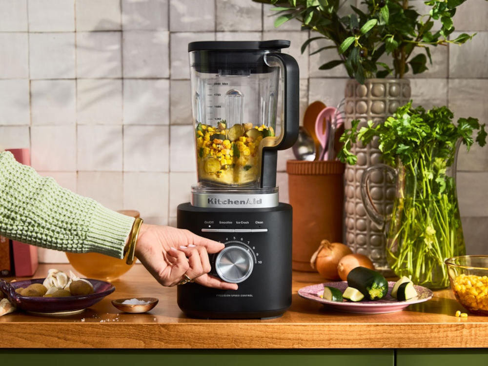 Na blacie kuchennym stoi czarny blender kielichowy z logo KitchenAid, wypełniony mieszanką warzyw. Obok znajdują się różne składniki, a ręka reguluje pokrętło na blenderze.