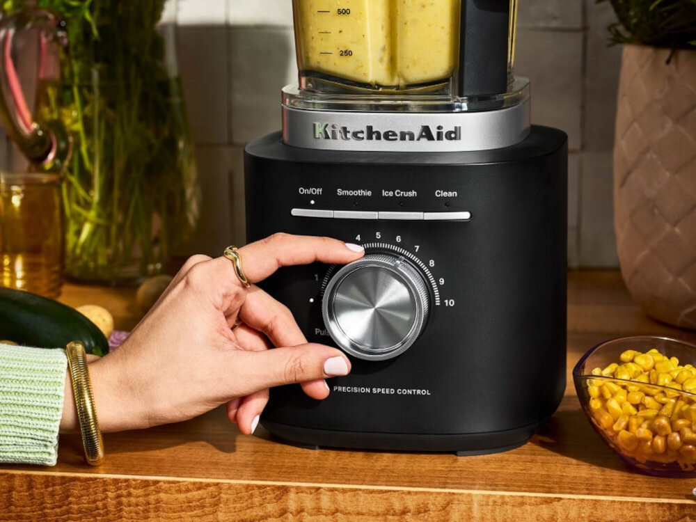 Czarny blender kielichowy z logo \'KitchenAid\' na froncie, z pokrętłem do regulacji prędkości i przyciskami oznaczonymi jako On/Off, Smoothie, Ice Crush, Clean. Obok blendera znajduje się miska z kukurydzą i inne kuchenne przedmioty.