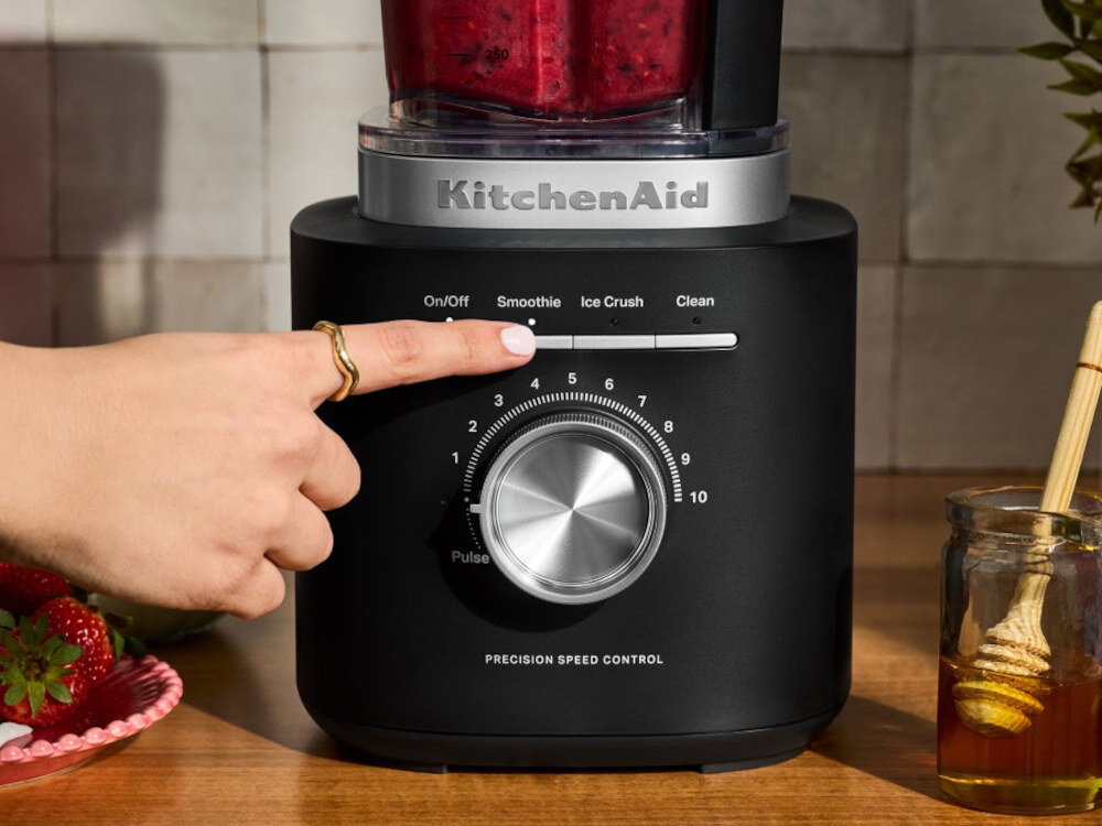 Czarny blender kielichowy z logo \'KitchenAid\' na górze, z pokrętłem do regulacji prędkości i przyciskami oznaczonymi jako On/Off, Smoothie, Ice Crush, Clean. Obok znajduje się słoik z miodem i truskawki na talerzu.