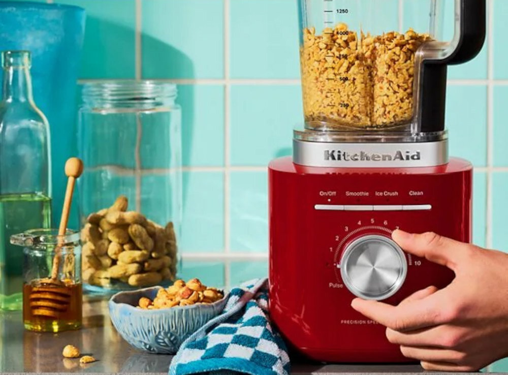 Czerwony blender kielichowy z logo \'KitchenAid\' na blacie kuchennym, wypełniony płatkami zbożowymi. Obok znajdują się słoiki z orzechami i miodem oraz niebieska ściereczka.