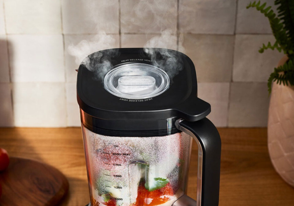 Na blacie kuchennym stoi czarny blender kielichowy z przezroczystym pojemnikiem, w którym znajdują się składniki do miksowania. Na pokrywie widoczny jest napis \'HEAT RELEASE VENT\', a z otworu unosi się para.