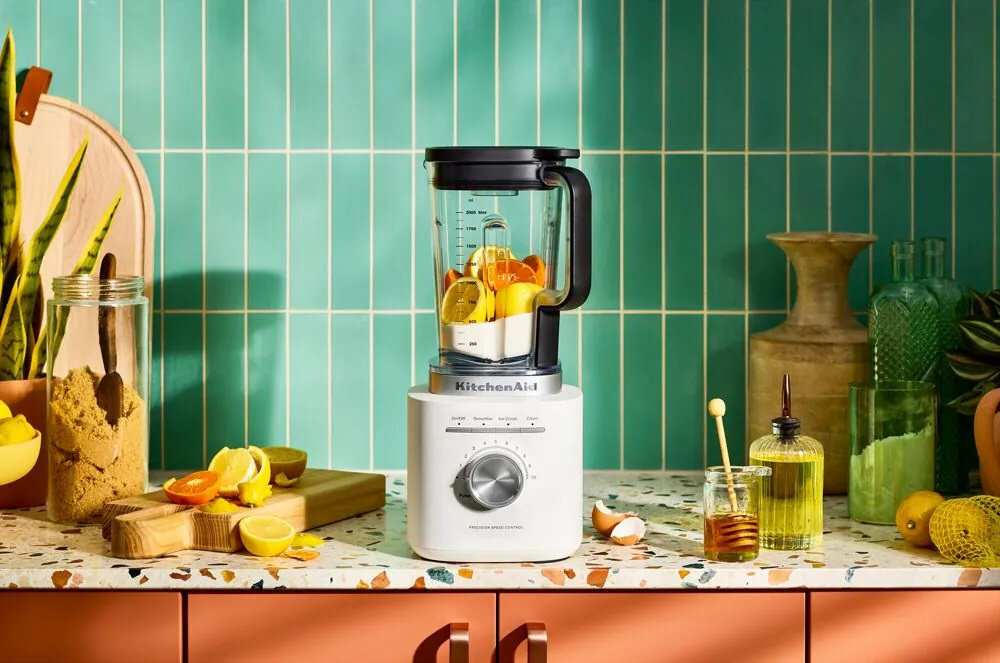 Na blacie kuchennym stoi blender kielichowy z logo KitchenAid, wypełniony kawałkami owoców. Wokół znajdują się różne akcesoria kuchenne, takie jak deska do krojenia z owocami i słoik z miodem.