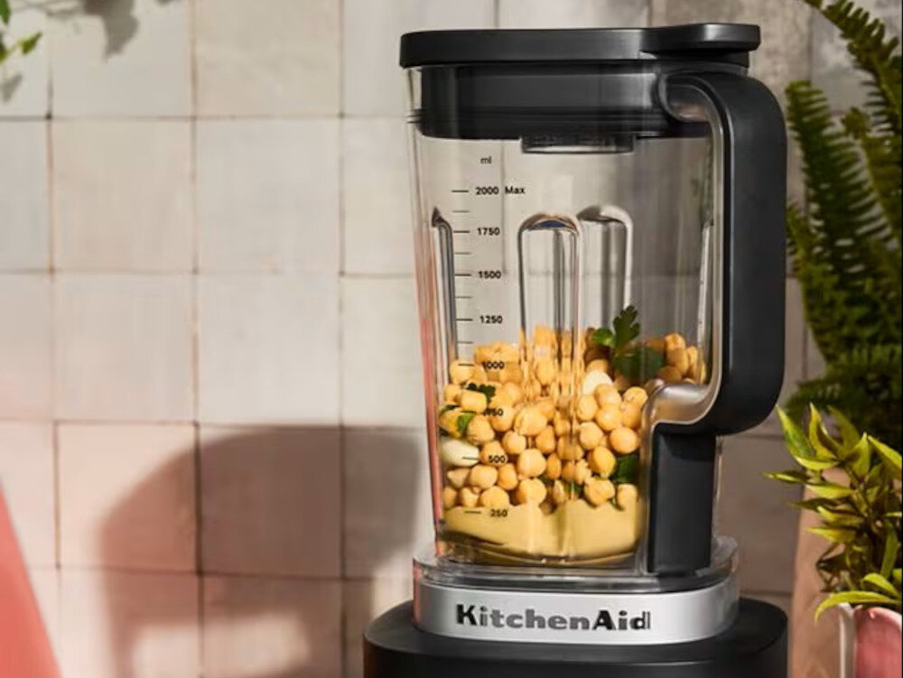 Blender kielichowy z logo KitchenAid, wypełniony ciecierzycą i ziołami, stoi na tle jasnych płytek ściennych. Na kielichu widoczna jest miarka w mililitrach do 2000 ml.