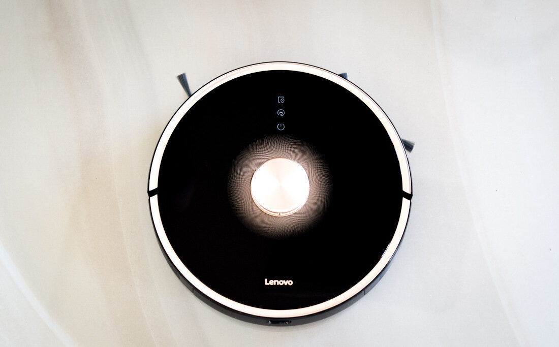 Czarny, okrągły robot sprzątający z widocznym logo Lenovo na górze. Na powierzchni znajdują się trzy ikony wskazujące funkcje urządzenia.