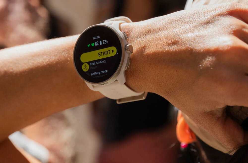 Osoba nosi smartwatch z białym paskiem, na ekranie widoczny jest tekst: \'14:26, 82 I 22h, START, Trail running, Basic, Battery mode\'.