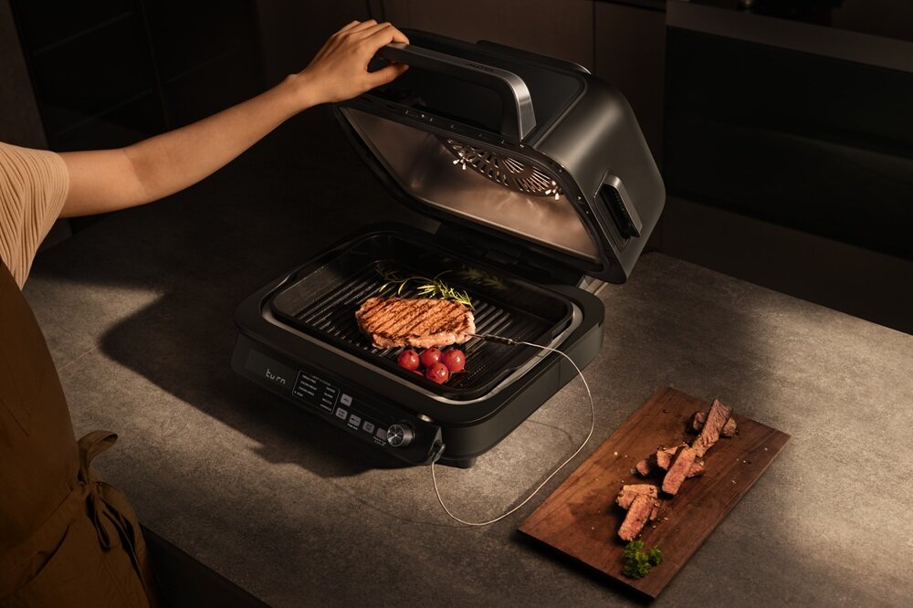 Osoba otwiera grill elektryczny, na którym znajduje się stek z ziołami i pomidorkami. Obok na desce leżą pokrojone kawałki mięsa.
