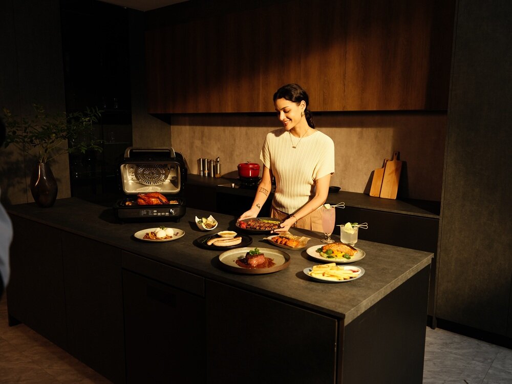 Kobieta stoi przy blacie kuchennym, na którym znajduje się grill elektryczny z otwartą pokrywą oraz kilka talerzy z różnymi potrawami. W tle widoczne są drewniane szafki i akcesoria kuchenne.