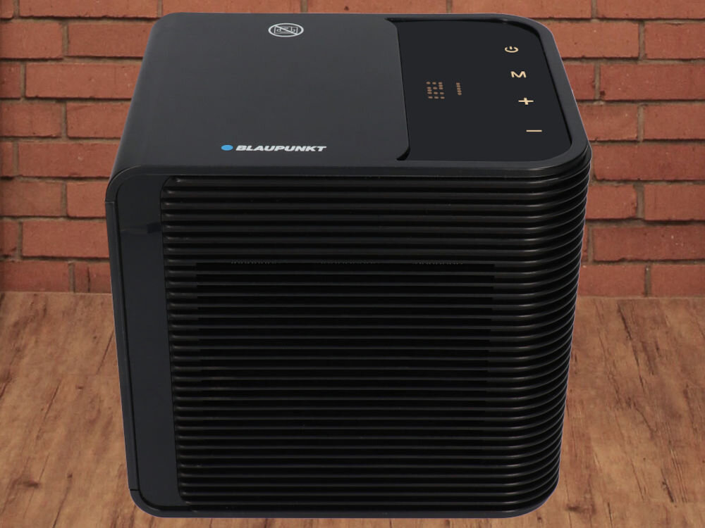 Czarny termowentylator z logo BLAUPUNKT na górze, posiada panel sterowania z symbolami i przyciskami. Urządzenie stoi na drewnianej podłodze, a w tle widoczna jest ceglana ściana.