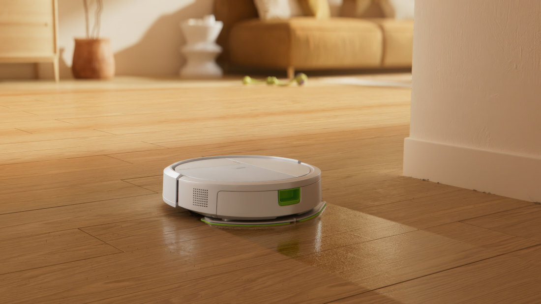 Na drewnianej podłodze znajduje się biały robot sprzątający. W tle widoczna jest część salonu z sofą i dekoracjami.