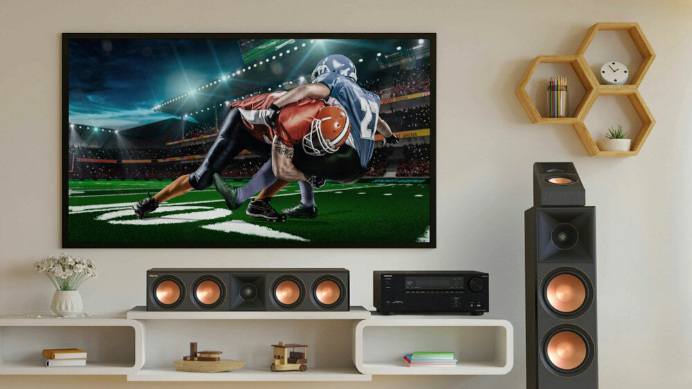 Na ścianie wisi telewizor wyświetlający scenę z meczu futbolu amerykańskiego. Na półce poniżej znajdują się głośniki i amplituner, a obok stoi duży głośnik podłogowy.