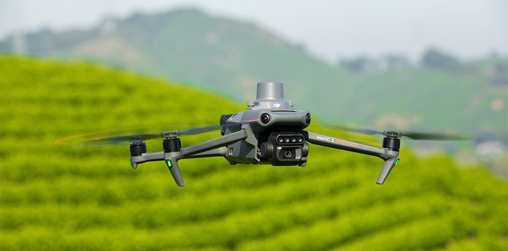 Dron z widoczną kamerą multispektralną unosi się nad zielonymi polami. Na dronie widoczny jest napis \'Mavic 3\'.