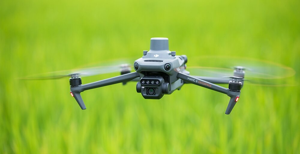 Dron z widoczną kamerą multispektralną unosi się nad zielonym polem. Na dronie widoczne jest logo DJI oraz napis \'Mavic 3 Multispectral\'.