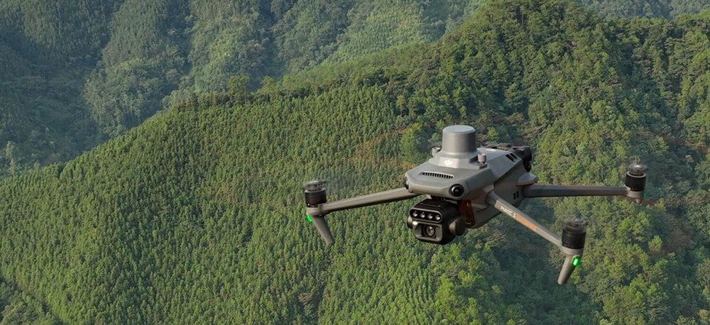 Dron z czterema wirnikami unosi się nad zalesionym terenem. Urządzenie ma zamontowaną kamerę i jest w kolorze szarym.