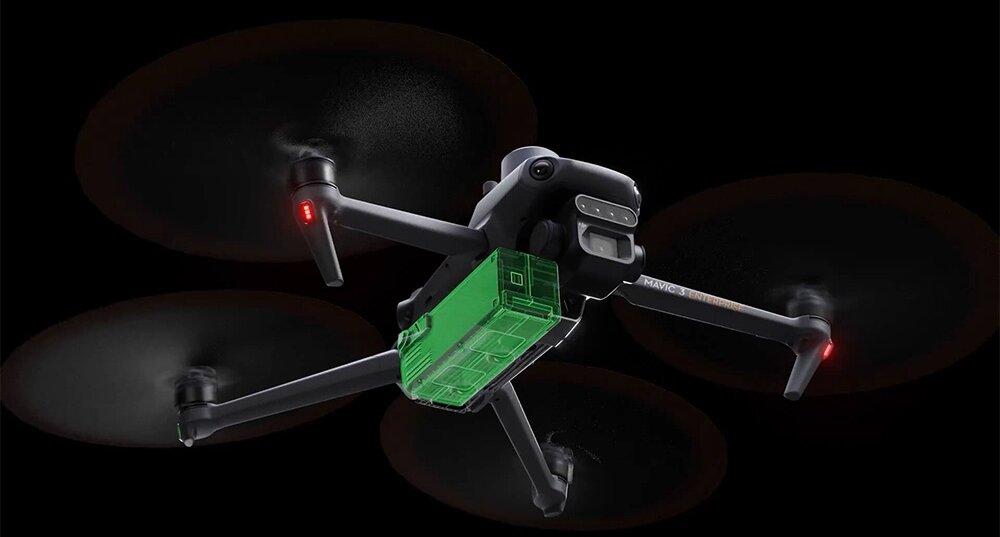 Na obrazie widoczny jest dron z czterema wirnikami, z zielonym elementem na spodzie. Dron unosi się w powietrzu na ciemnym tle.