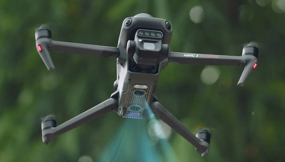 Dron w locie z widocznymi czterema ramionami i kamerą na spodzie. Na ramieniu napis \'MAVIC 3 Multispectral\'.
