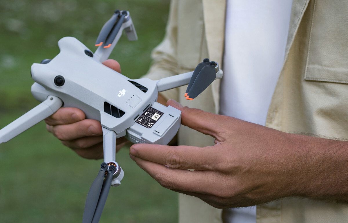 Osoba trzyma drona z widocznym logo \'dji\', wkładając lub wyjmując baterię. Dron ma szare ramiona i pomarańczowe końcówki śmigieł.