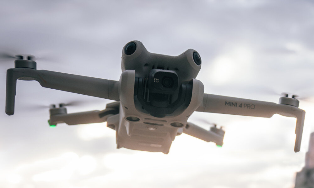 Dron w locie na tle pochmurnego nieba, z widocznym napisem \'MINI 4 PRO\' na ramieniu. Dron ma cztery śmigła i kamerę z przodu.
