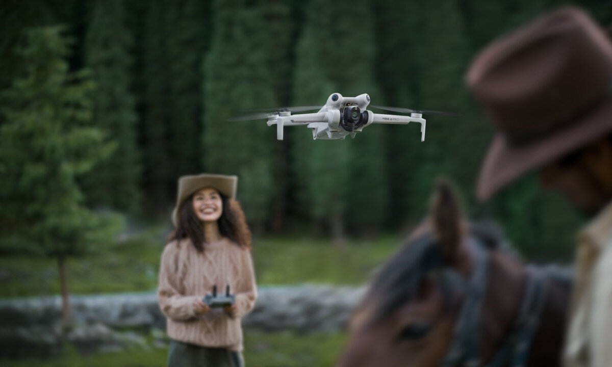 Dron unosi się w powietrzu na tle zalesionego terenu. W tle widoczne są osoby, jedna z nich trzyma kontroler.