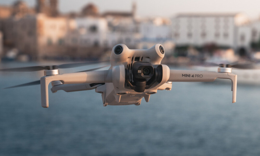 Dron w kolorze jasnym unosi się nad wodą, w tle widać rozmyte budynki. Na ramieniu drona widoczny jest napis \'MINI 4 PRO\'.