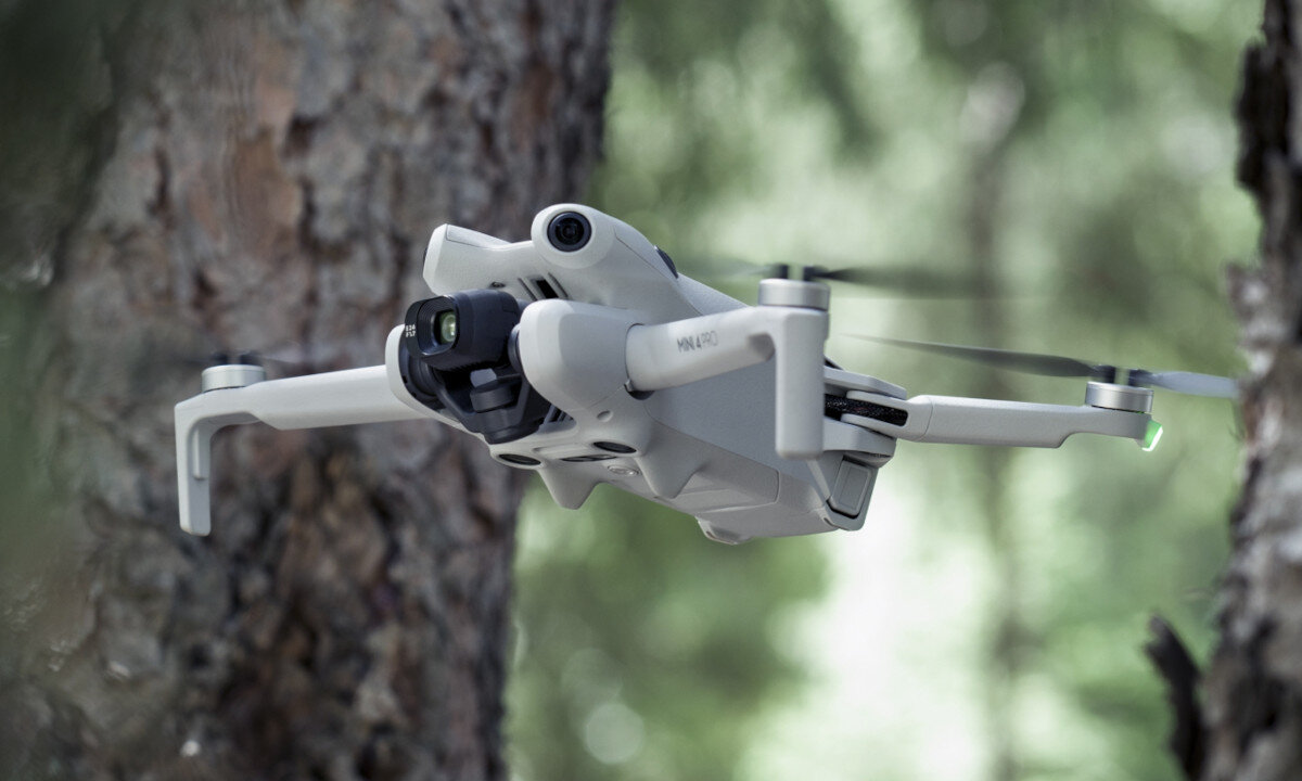 Dron w kolorze szarym unosi się w powietrzu na tle drzew. Widoczne są jego śmigła oraz kamera z przodu.