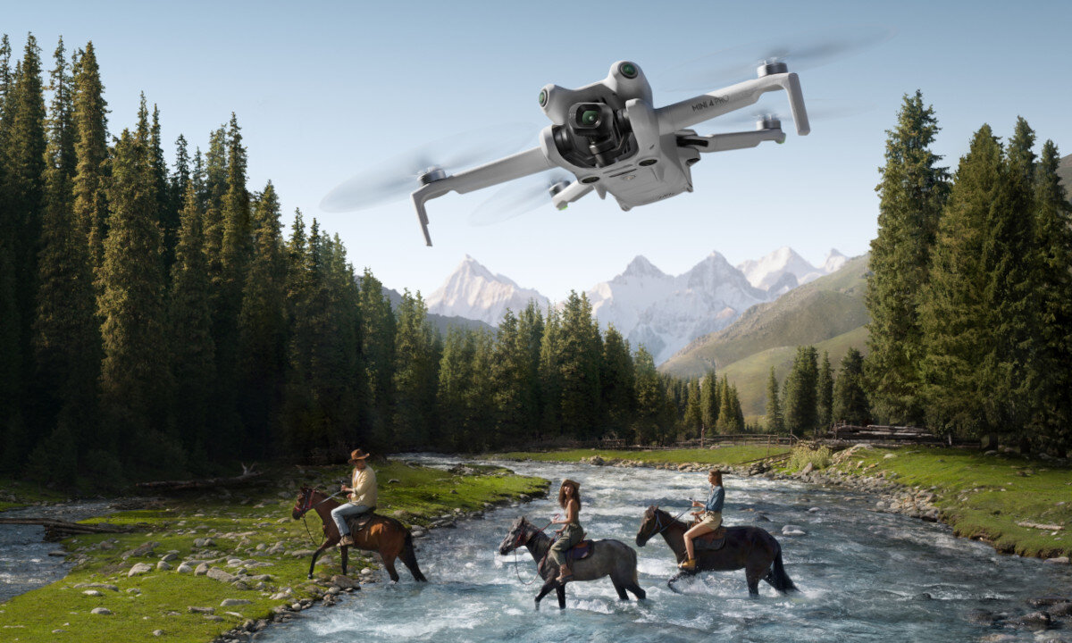 Dron unosi się nad rzeką, nad którą trzech jeźdźców na koniach przeprawia się przez wodę. W tle widoczne są góry i lasy.