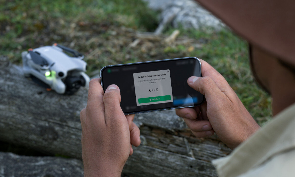 Osoba trzyma smartfon z wyświetlonym komunikatem \'Switch to QuickTransfer Mode\'. Obok na drewnianym pniu leży mały dron z zielonym światłem.