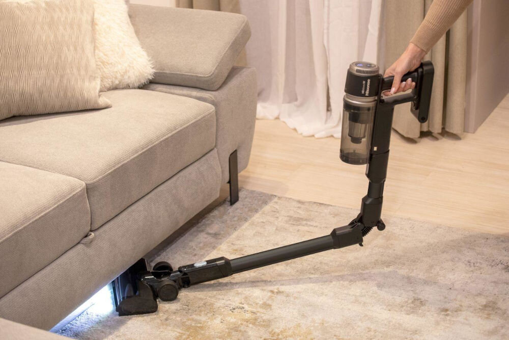 Osoba używa odkurzacza pionowego z elastyczną rurą, aby odkurzyć przestrzeń pod kanapą. Odkurzacz jest bezprzewodowy i ma przezroczysty pojemnik na kurz.