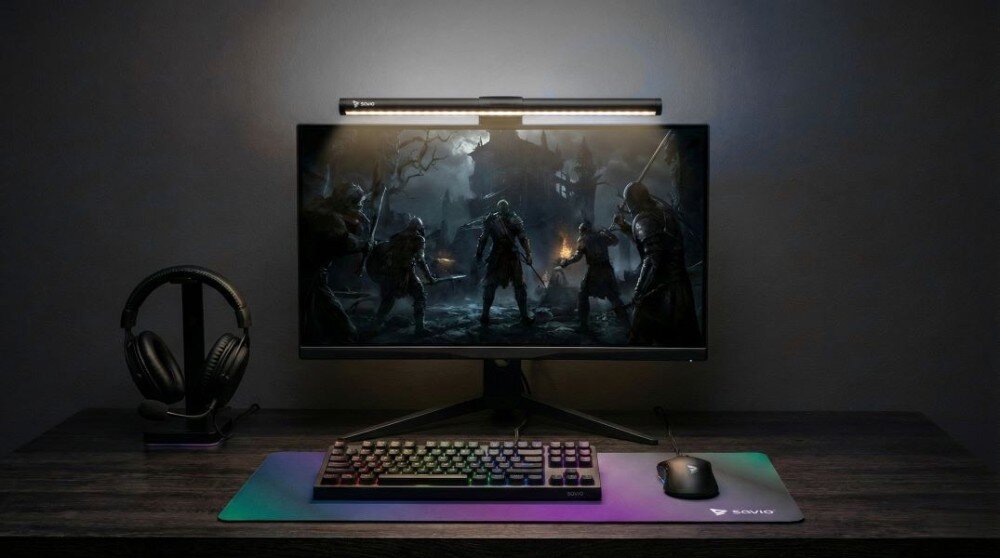 Na biurku znajduje się monitor z włączoną grą, nad którym zamontowana jest lampka na monitor. Obok monitora leżą słuchawki, klawiatura i myszka na podkładce z logo \'SAVIO\'.