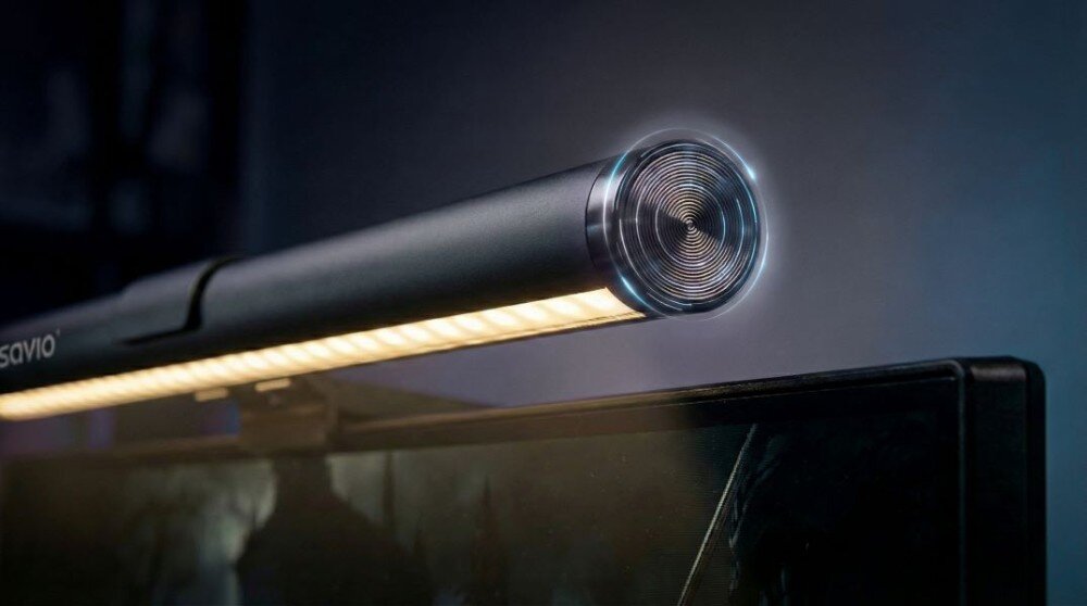 Lampka na monitor o cylindrycznym kształcie z widocznymi diodami LED, zamontowana na górnej krawędzi ekranu. Na obudowie widoczne jest logo \'savio\'.