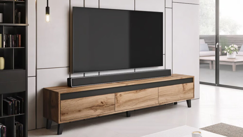 Na obrazie widoczna jest szafka RTV w kolorze dębu z trzema szufladami, na której stoi duży telewizor i soundbar. W tle znajduje się nowoczesne wnętrze z dużym oknem i półką z książkami.