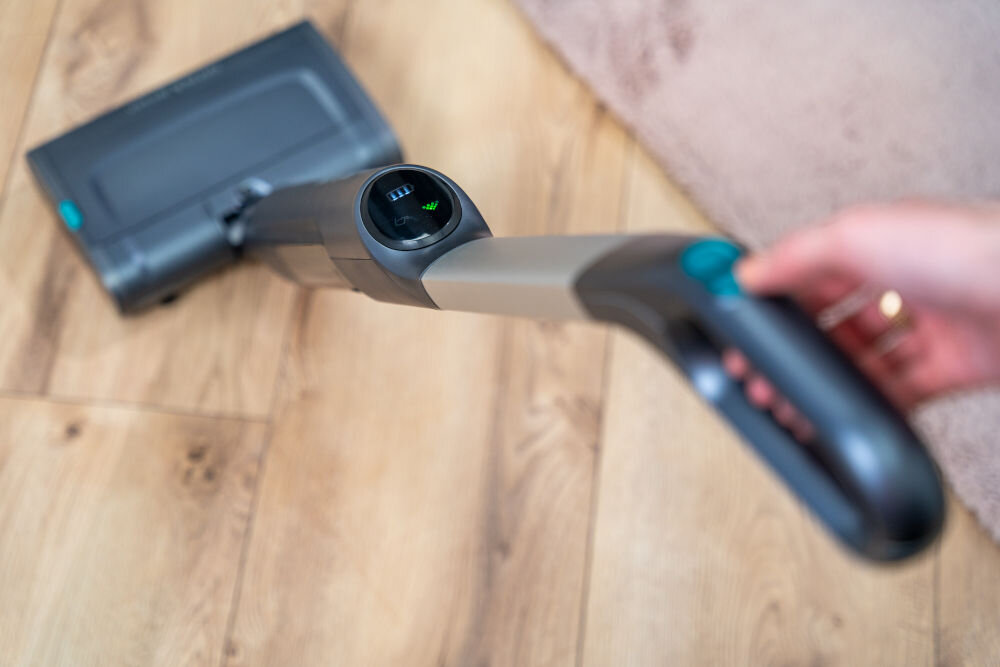 Osoba trzyma bezprzewodowy mop elektryczny z wyświetlaczem pokazującym poziom naładowania baterii. Urządzenie znajduje się na drewnianej podłodze obok dywanu.