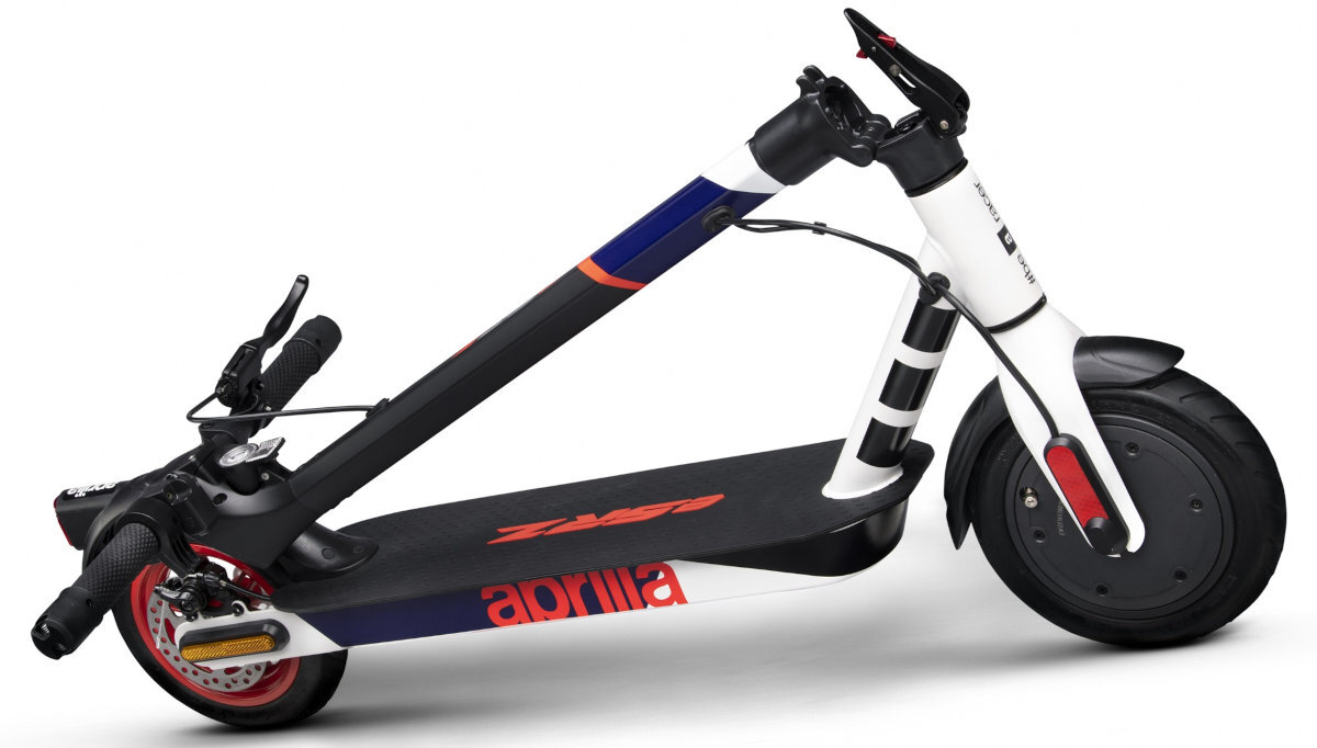 Hulajnoga elektryczna w kolorach biało-czarno-czerwonym z widocznym logo \'aprilia\' na podeście. Posiada duże koła, kierownicę z hamulcami i składany mechanizm.