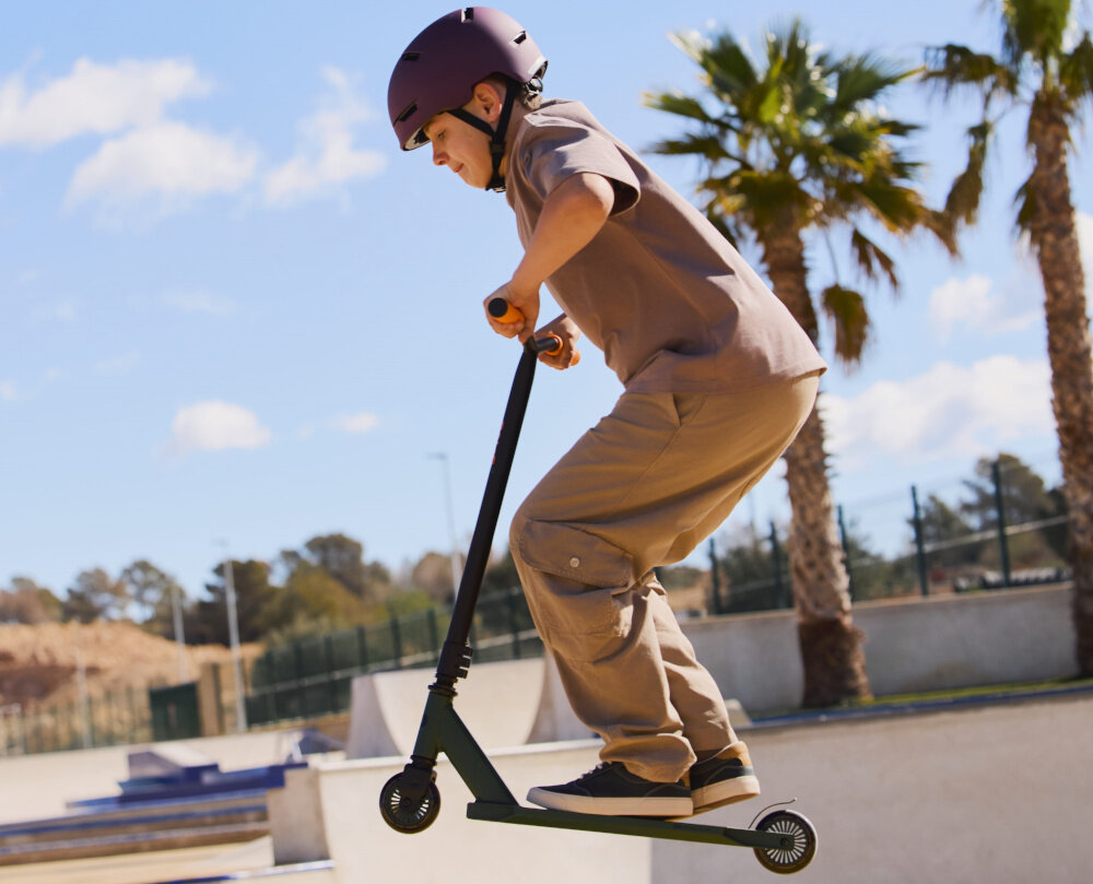 Osoba w kasku wykonuje skok na zielonej hulajnodze wyczynowej na skateparku. W tle widoczne są palmy i niebo z chmurami.