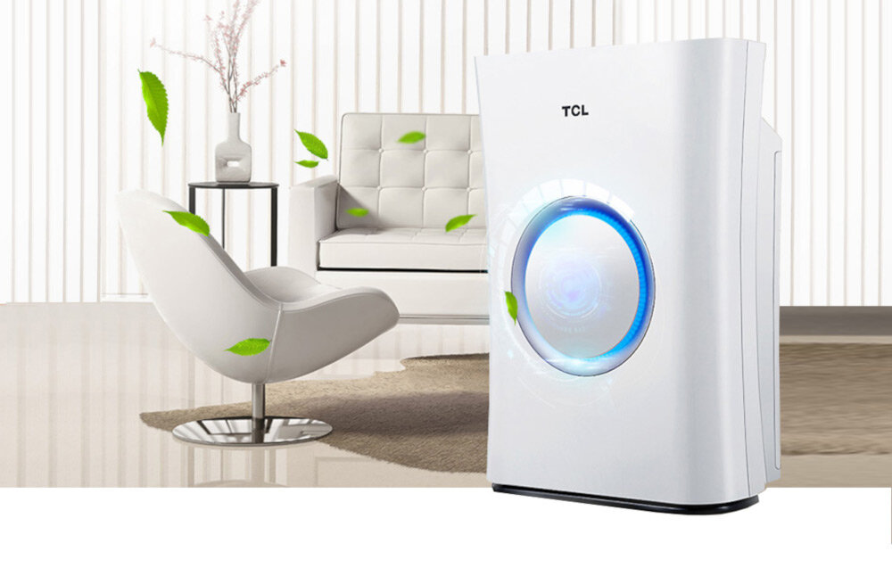 Na obrazie znajduje się biały oczyszczacz powietrza z logo TCL, obok nowoczesnego fotela i stolika z wazonem. W tle widoczna jest sofa, a wokół unoszą się zielone liście.