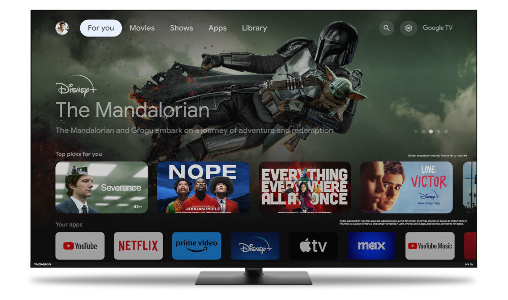 Telewizor wyświetla ekran główny z aplikacjami takimi jak YouTube, Netflix, Prime Video, Disney+, Apple TV, Max i YouTube Music. Na ekranie widoczny jest także baner promujący serial \'The Mandalorian\' na Disney+.