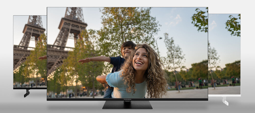 Na ekranie telewizora wyświetlany jest obraz kobiety z dzieckiem na tle wieży Eiffla i drzew. Telewizor ma cienką ramkę i stoi na prostokątnej podstawie.