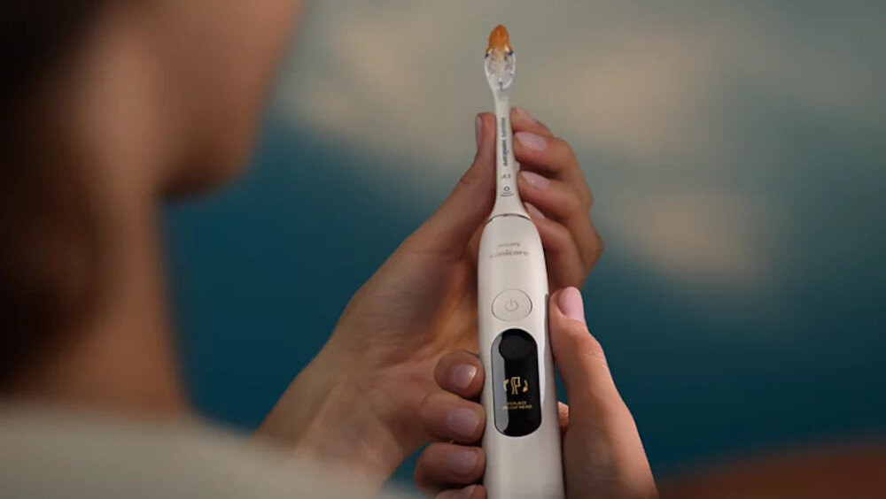 Osoba trzyma białą szczoteczkę soniczną z widocznym przyciskiem i wyświetlaczem. Na szczoteczce znajduje się logo \'PHILIPS\'.