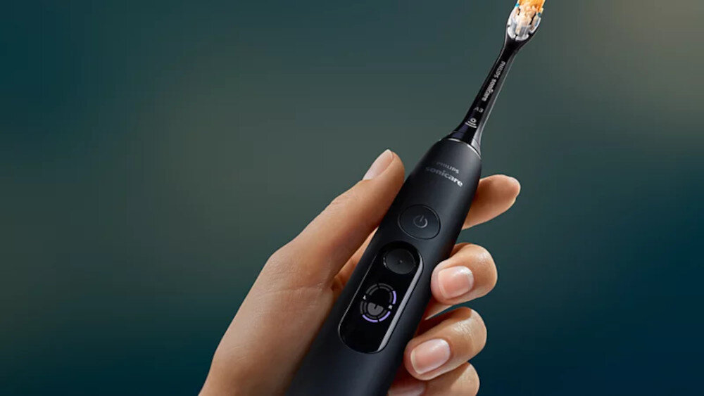 Osoba trzyma czarną szczoteczkę soniczną z widocznymi przyciskami i podświetleniem. Na szczoteczce widoczny jest napis \'sonicare\'.