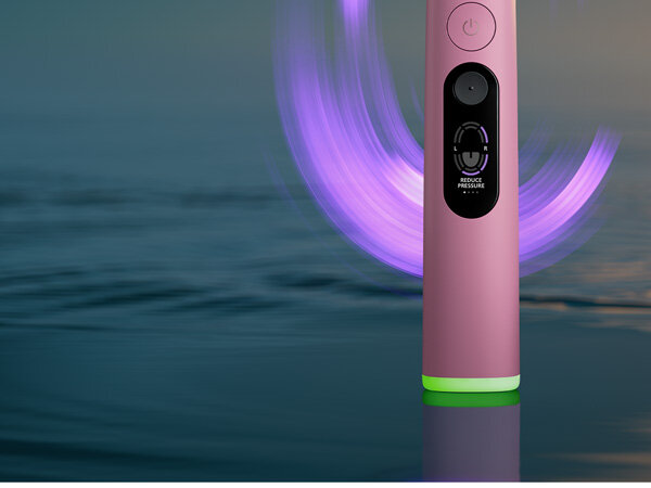 Różowa szczoteczka soniczna z wyświetlaczem pokazującym napis \'REDUCE PRESSURE\'. W tle widoczna jest rozmyta powierzchnia wody i fioletowe światło.