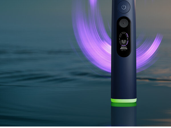 Granatowa szczoteczka soniczna z podświetleniem w kolorze fioletowym, widoczny przycisk zasilania oraz napis \'REDUCE PRESSURE\'.